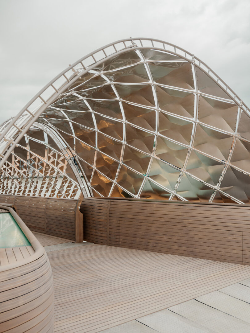 Curved metallic structure with a lattice design sits behind wooden decking and paneling under a cloudy sky.