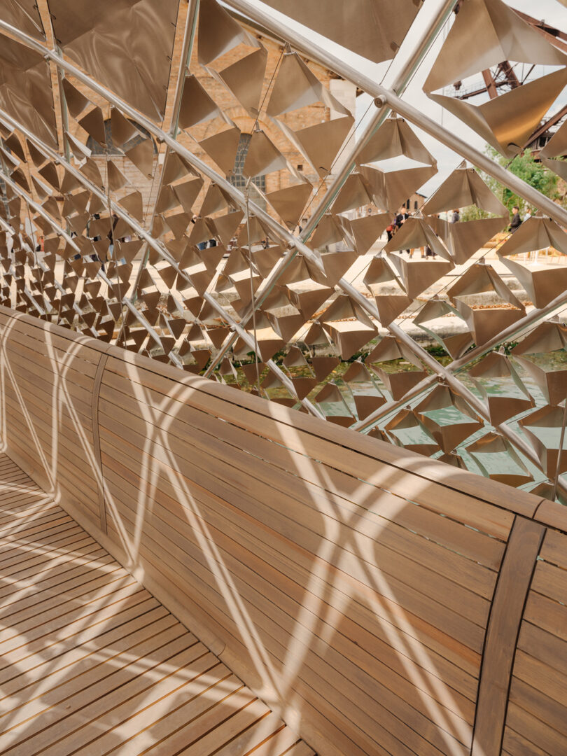 Wooden bench under a geometric metal lattice structure that casts patterned shadows onto the seating and floor.