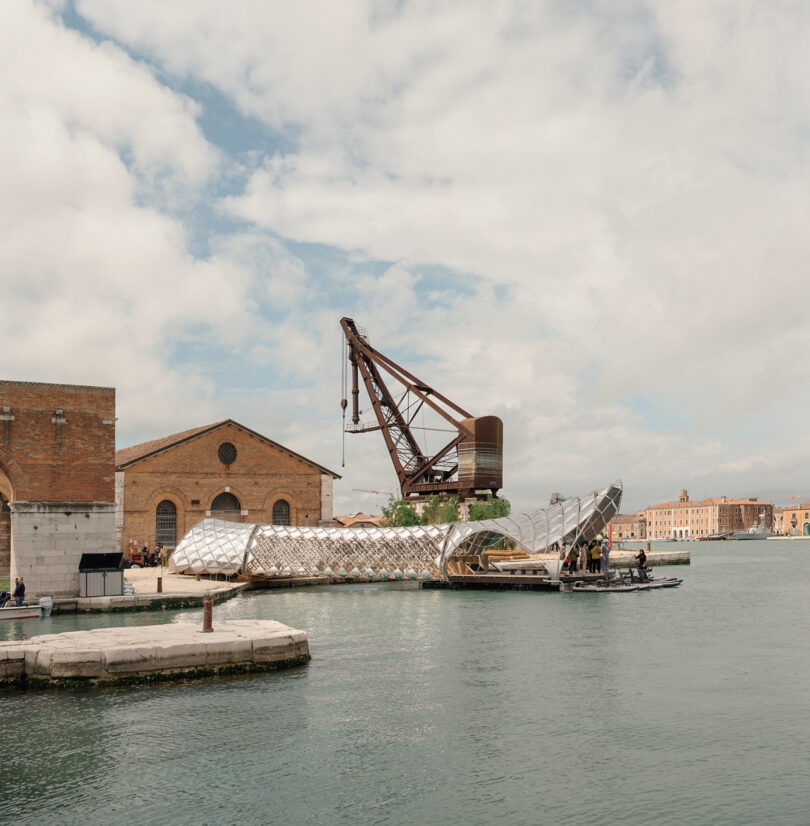 A large industrial crane stands beside a brick warehouse at a waterfront, with a modern, metallic structure and people near the water under a partly cloudy sky.