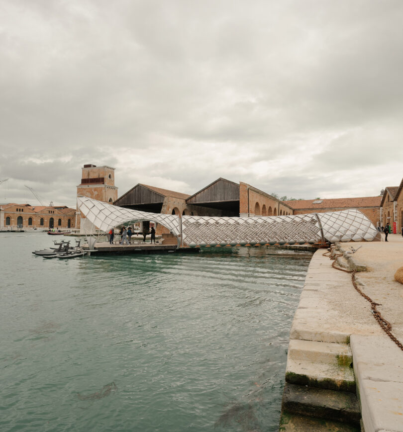A modern, lattice-structured pavilion extends over the water beside historic brick buildings on a cloudy day. Several people are gathered under the pavilion.