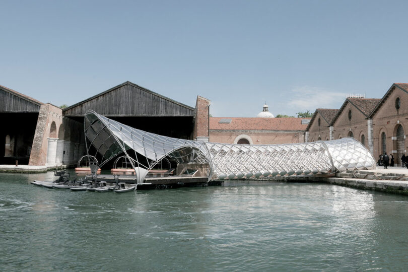 A futuristic, Porsche-inspired, lattice-structured pavilion designed by Norman Foster extends over the waterfront between historic industrial brick buildings under a clear sky.