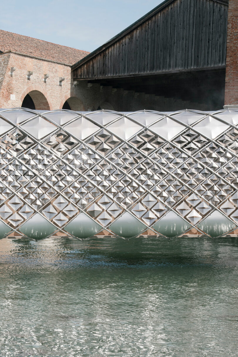 A geometric, lattice-patterned structure, designed by Norman Foster, floats on water in front of historic brick buildings with arched openings.