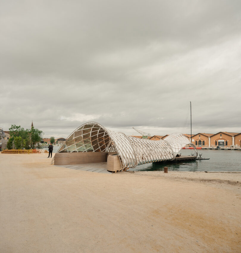 A modern, arched pavilion with a lattice roof extends from a sandy shore into the water, with a sailboat docked beside it under a cloudy sky.