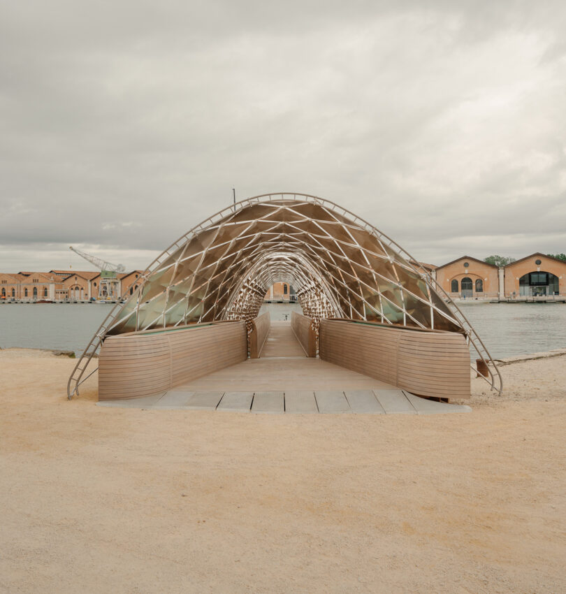 A modern arched pedestrian bridge with a lattice framework spans over water, with sandy ground in the foreground and industrial buildings in the background under a cloudy sky.