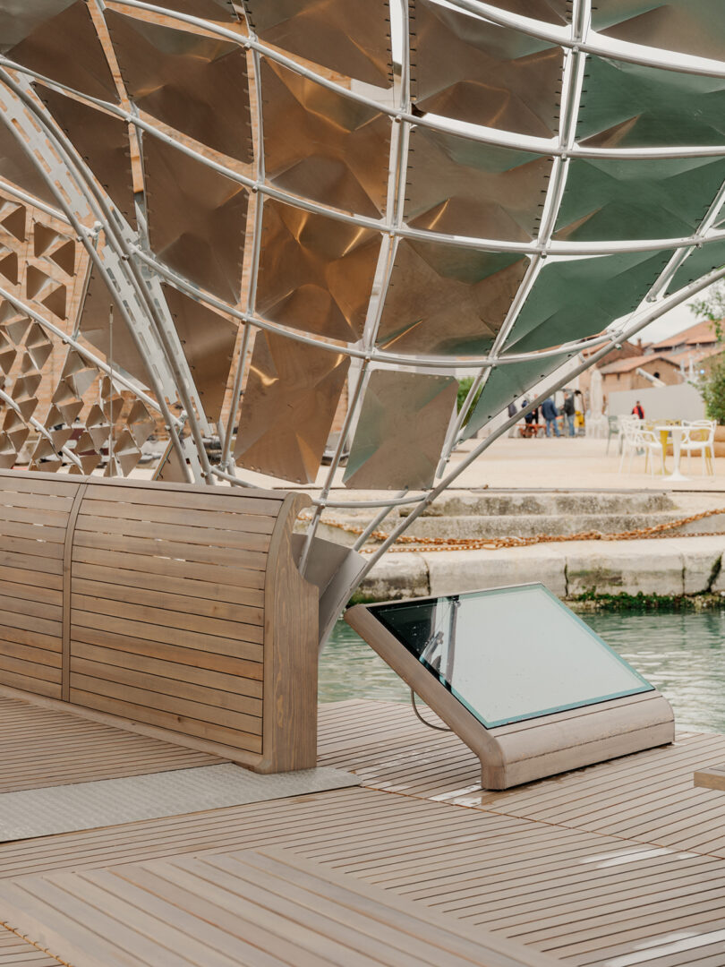 Curved wooden and metal structure with a partially open lattice canopy, adjacent to a body of water and an angled glass panel in the foreground.