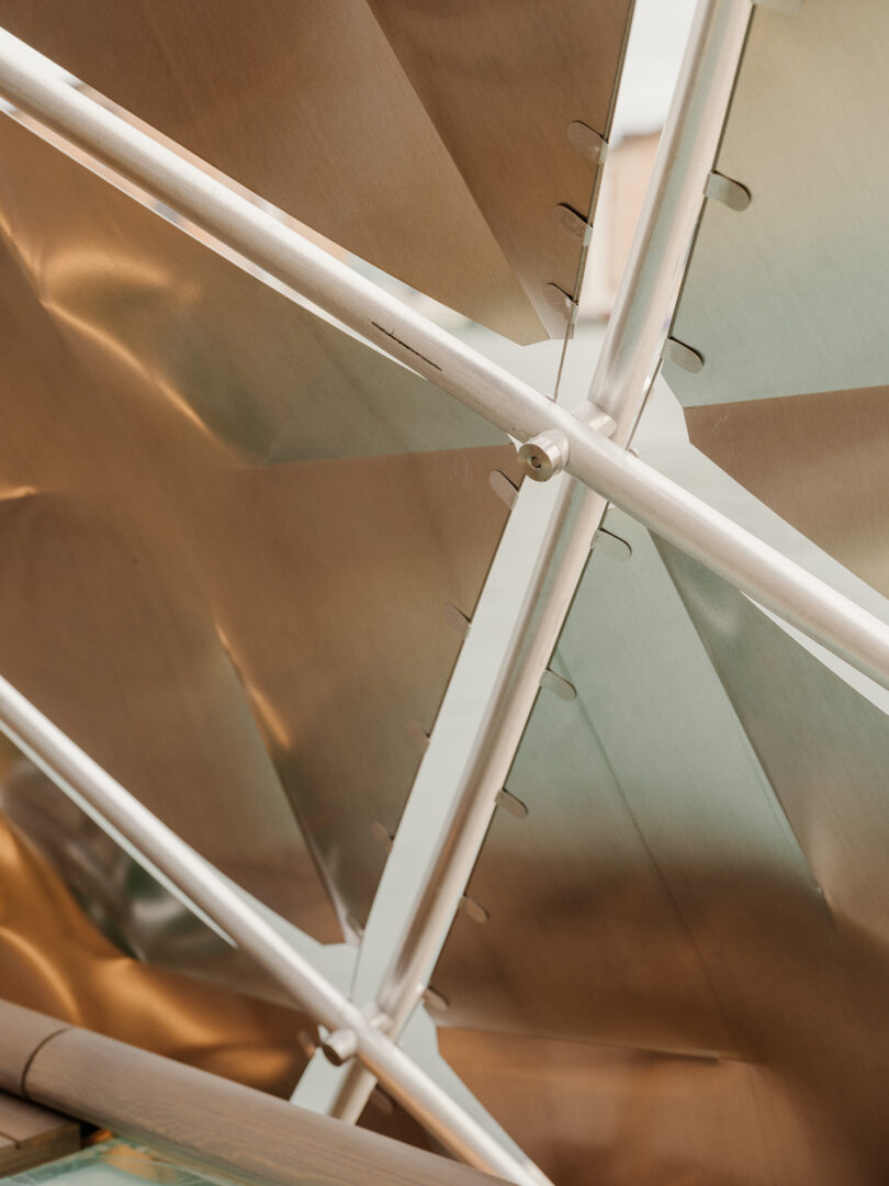 Close-up view of intersecting metal rods and angled metal panels, showing structural details and reflective surfaces in a modern architectural element.