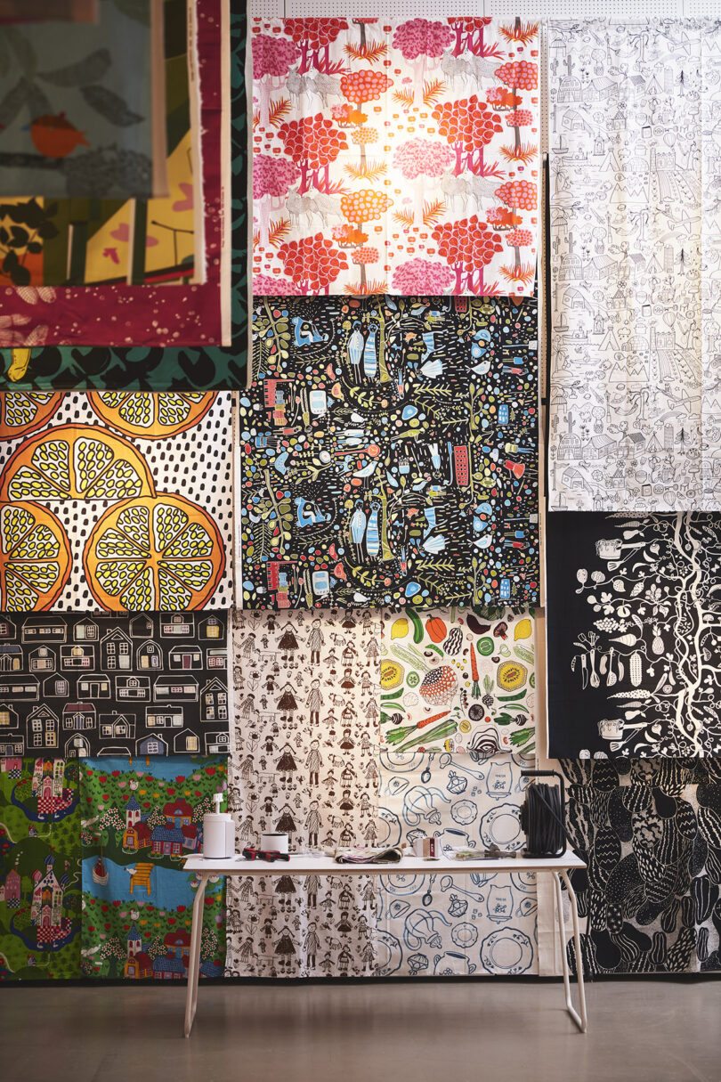 Colorful patterned fabrics are displayed hanging on a wall above a white table with a tissue box and a cup on it in a well-lit room.