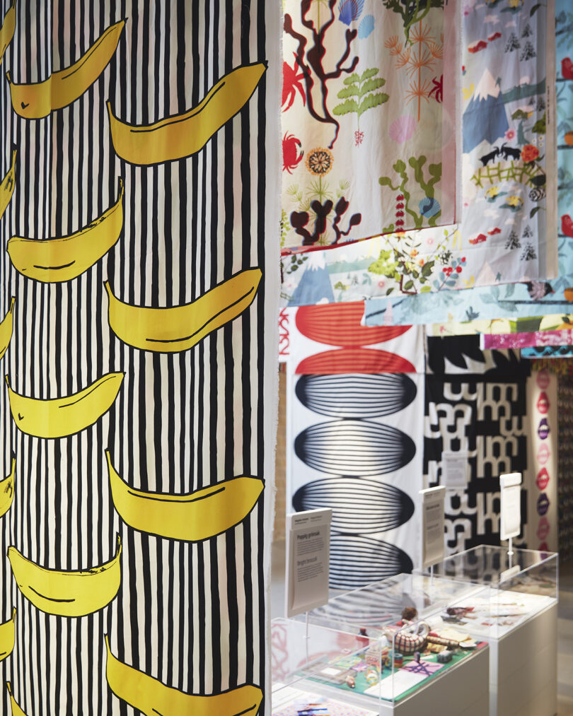 A display of colorful patterned textiles, including one with yellow bananas on black and white stripes, in a well-lit exhibition space.