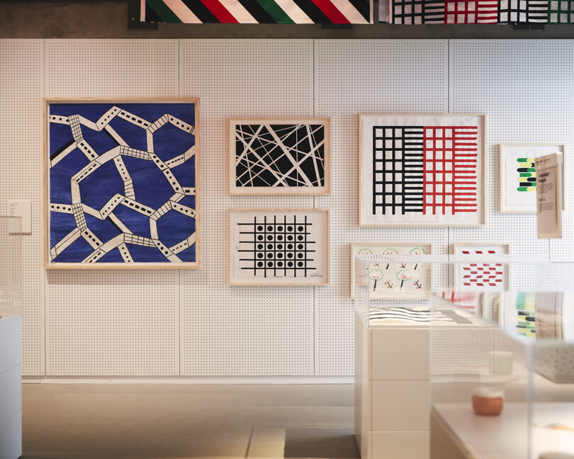 Framed abstract geometric artworks featuring grids, lines, and patterns hang on a white pegboard wall in a modern gallery space.