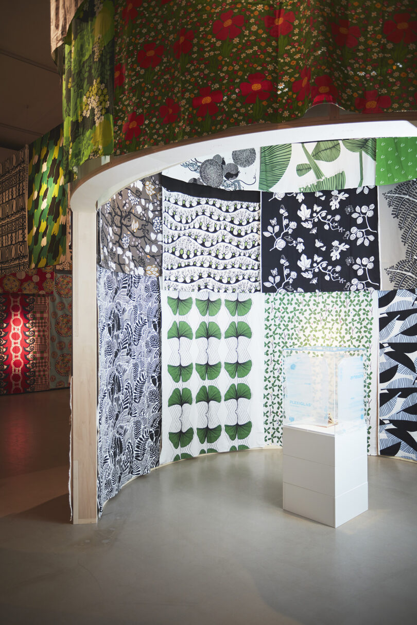 A display of patterned fabric panels in black, white, and green hangs around a curved wall; a small sculpture is showcased on a white pedestal in the foreground.