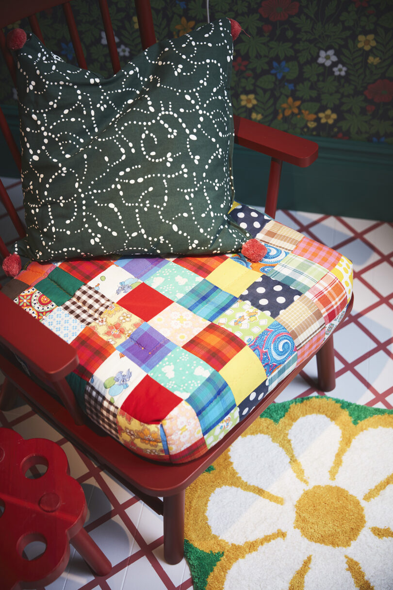 A wooden chair with a colorful patchwork cushion and a green pillow featuring a white dotted pattern, next to a daisy-patterned rug on a tiled floor.