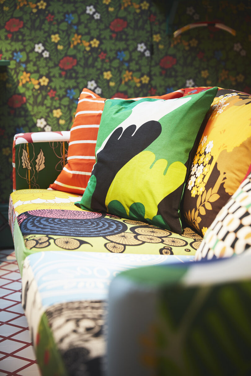 A sofa with colorful, patterned cushions and upholstery sits in a room with floral wallpaper and a tiled floor.