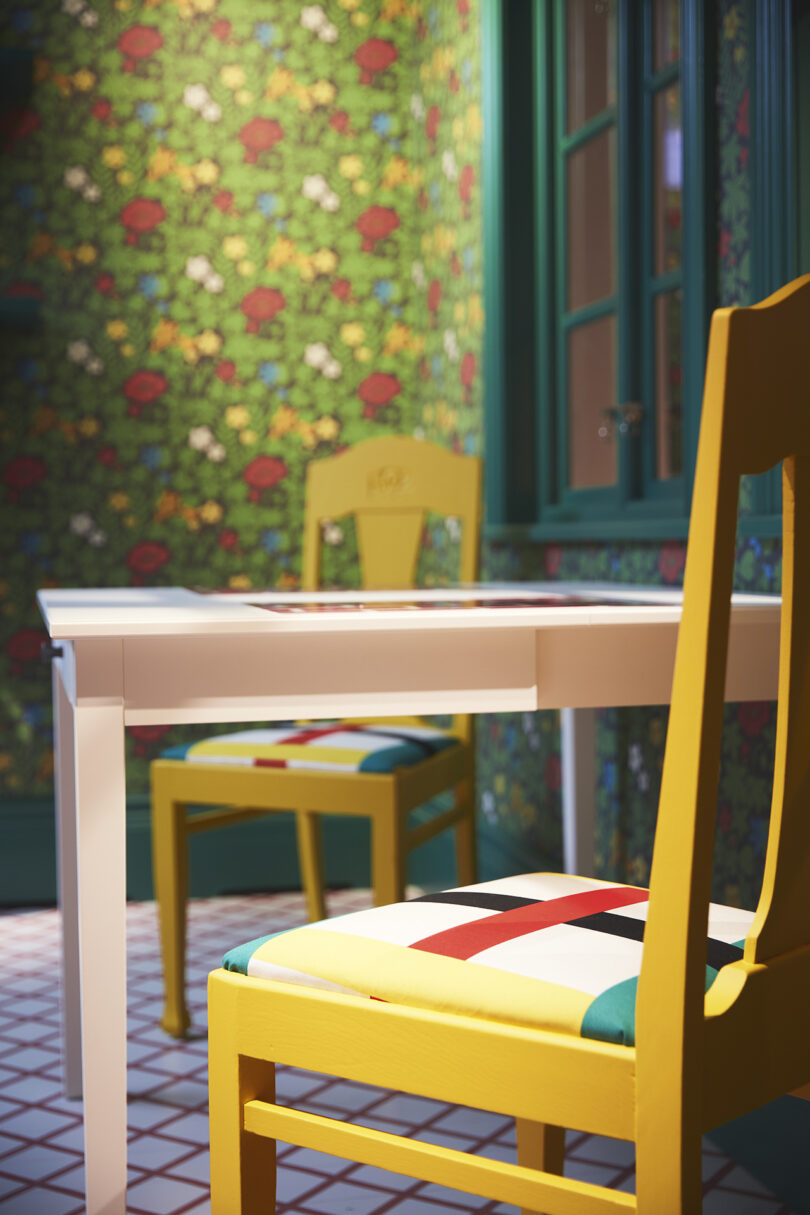 Two yellow chairs with patterned seats are positioned at a white table in a room with green floral wallpaper and tiled floor.