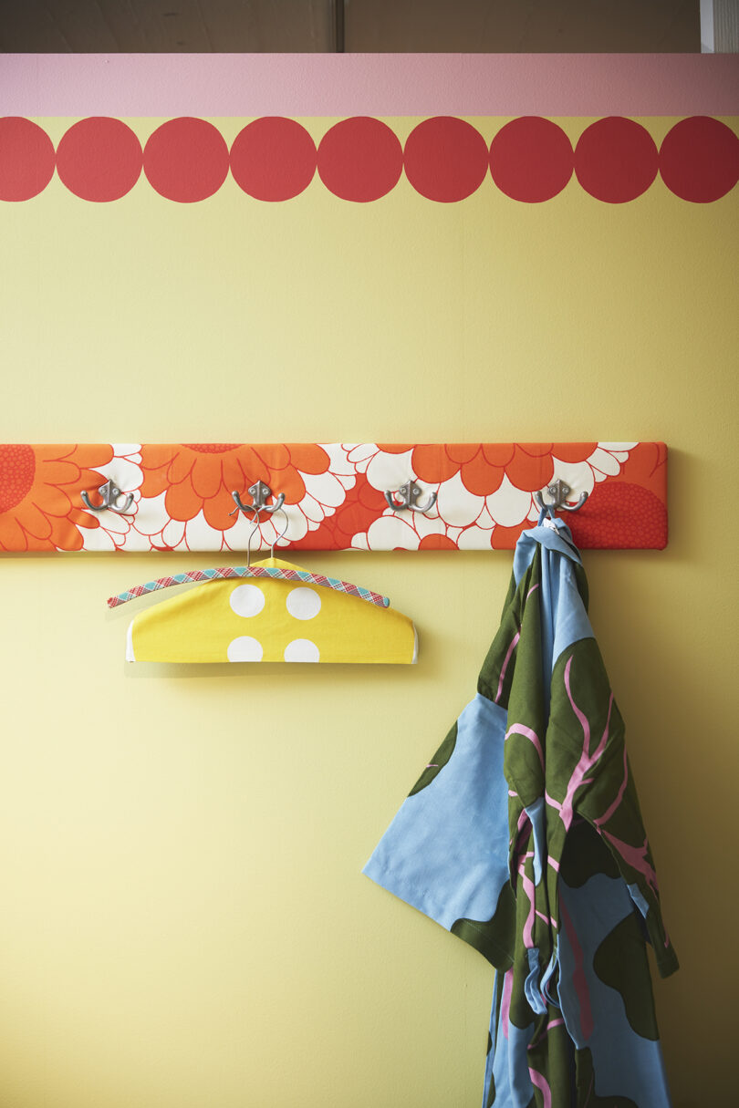 A yellow wall with a red floral coat rack holding two colorful garments, and a yellow polka-dot hanger suspended below the rack.