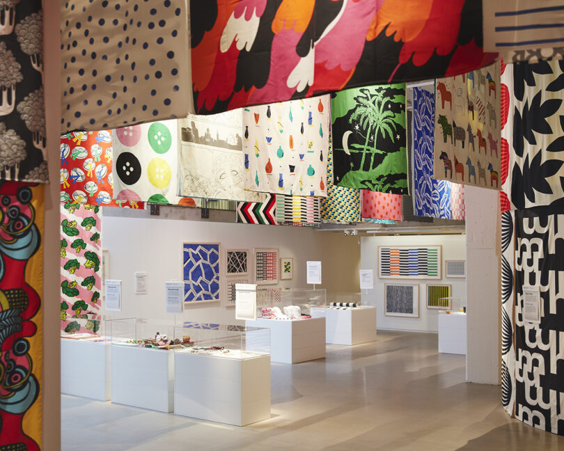 A bright exhibition space displays various colorful textiles hanging from the ceiling, with art pieces and informational panels arranged on white pedestals and walls.