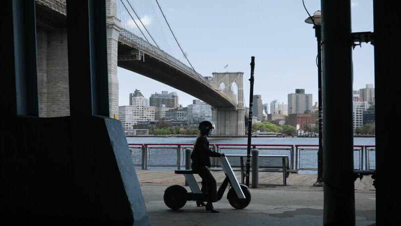 A person wearing a helmet rides an electric bike under a bridge by the waterfront, with city buildings visible in the background.
