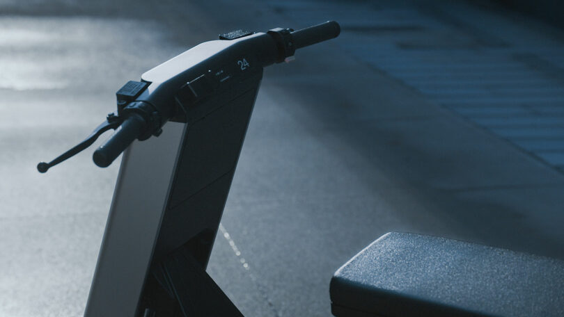 Close-up view of the handlebars and seat of an electric scooter, photographed outdoors on a paved surface in low light.