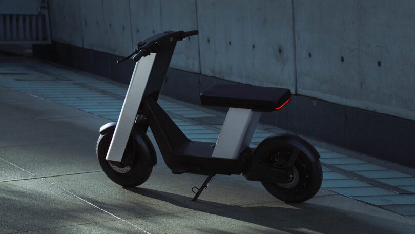 A modern electric scooter with a seat is parked on a sidewalk near a concrete wall, illuminated by soft ambient light.
