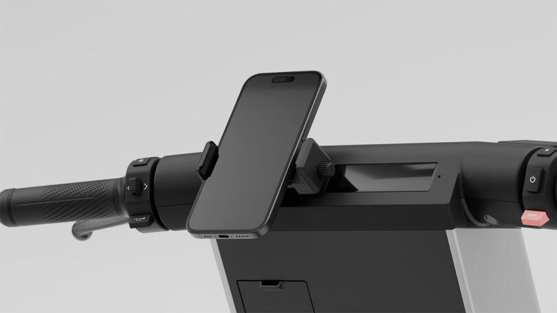 Close-up of a smartphone mounted on the handlebar of an electric scooter, with visible grip, control buttons, and part of the scooter body.