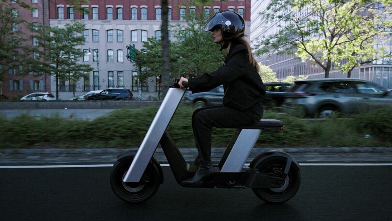 A person wearing a helmet rides a modern electric scooter on a city street with cars and buildings in the background.