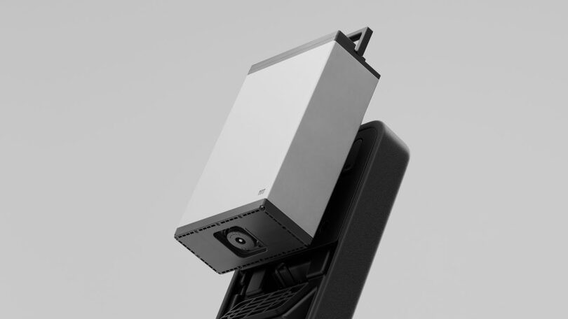 A close-up of a rectangular, metallic electronic device with a visible camera lens, partially inserted into a black docking station against a plain background.