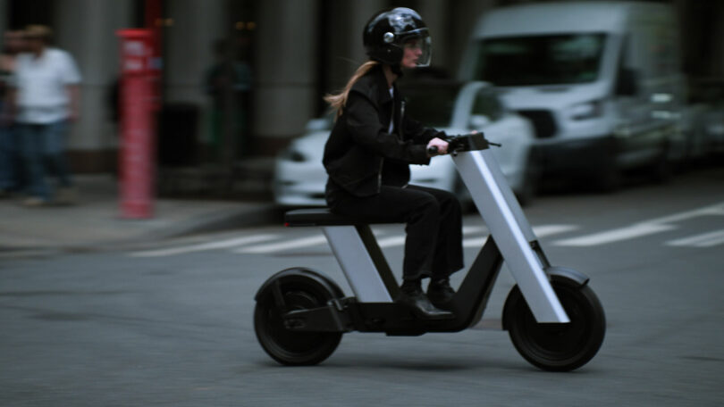 A person wearing a helmet rides an electric scooter on a city street, with parked cars and pedestrians visible in the blurred background.