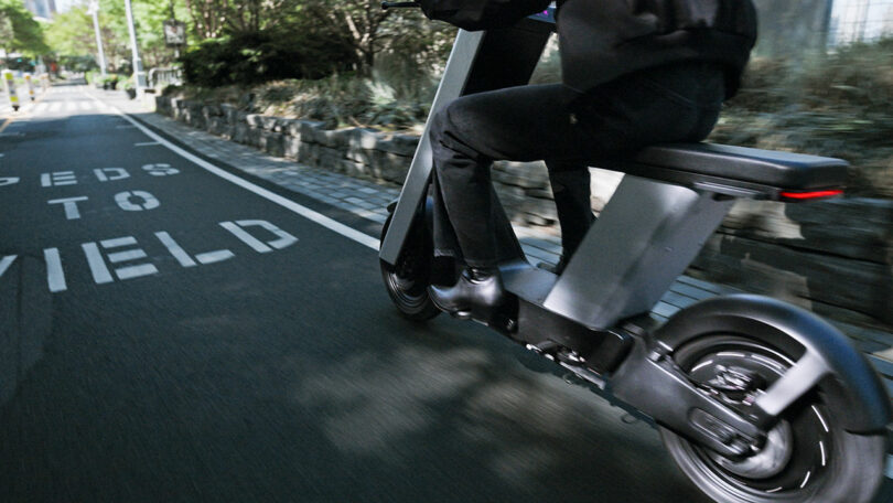 Person riding a black electric scooter on a paved path with "PEDS TO YIELD" painted on the ground and greenery on the side.