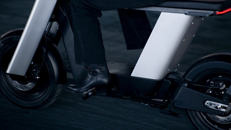A close-up of a person riding a modern electric scooter, focusing on the lower body, footrests, and wheels, with the vehicle in motion.