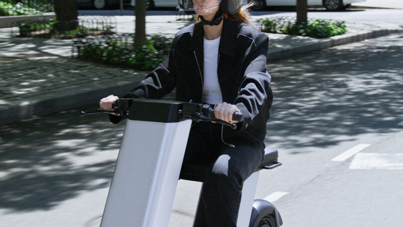 A person wearing a helmet and black jacket rides an electric scooter on a city street during the day.