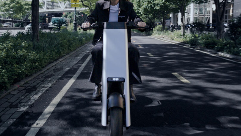 A person rides a modern electric bike on a designated bike lane in an urban outdoor setting with trees and buildings.