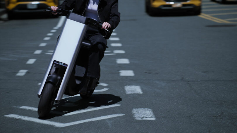 A person rides a modern electric scooter in a bike lane on a city street, with cars visible in the background.