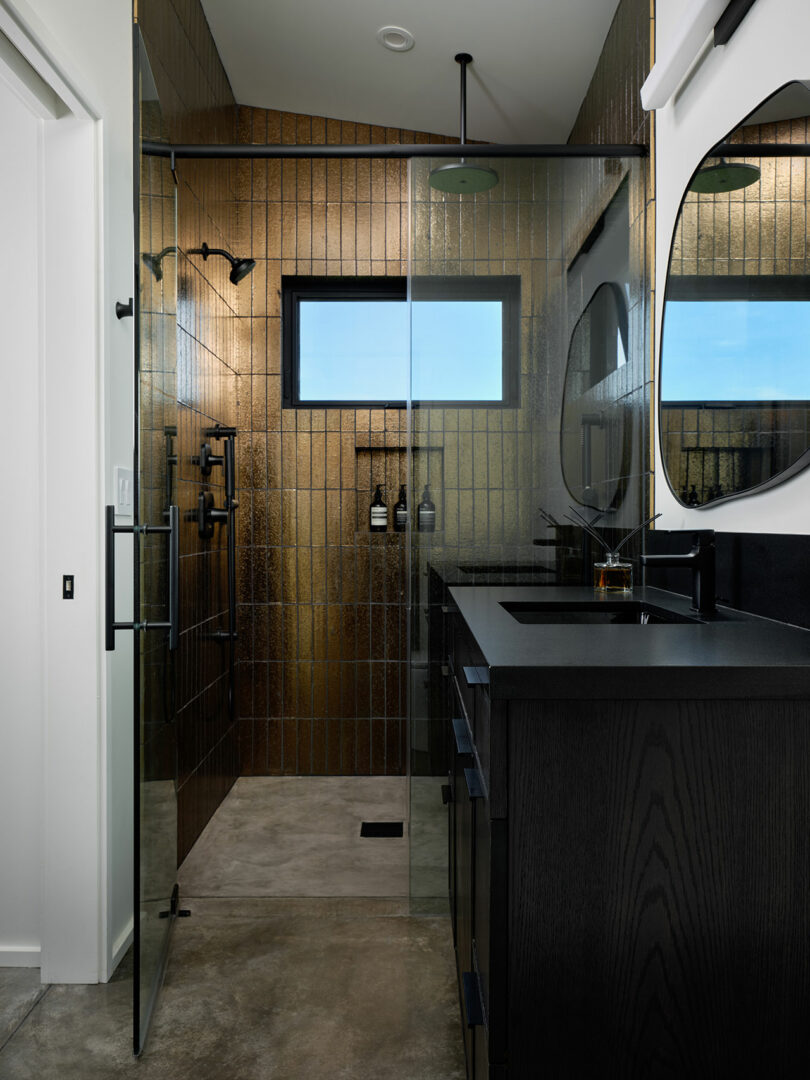 Modern bathroom with black fixtures, glass shower enclosure, textured metallic gold tile walls, a small window, and a black countertop with round mirror.