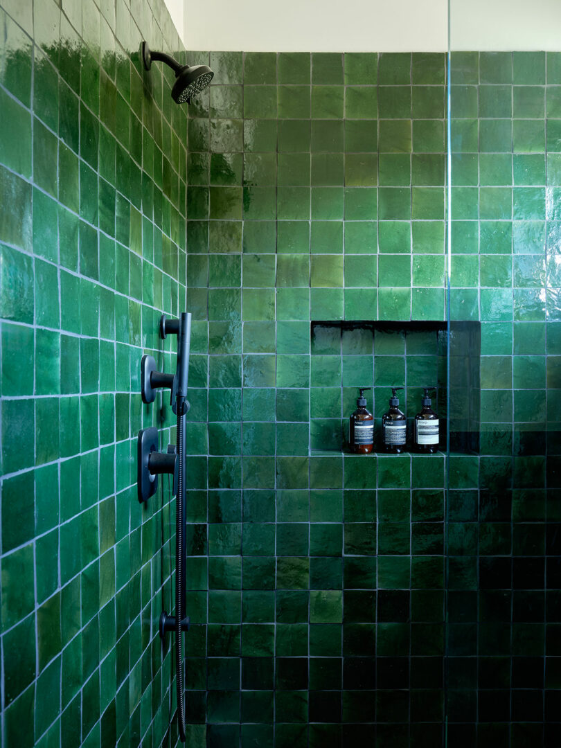 A shower with glossy green square tiles, matte black fixtures, and a recessed shelf holding three bottles.