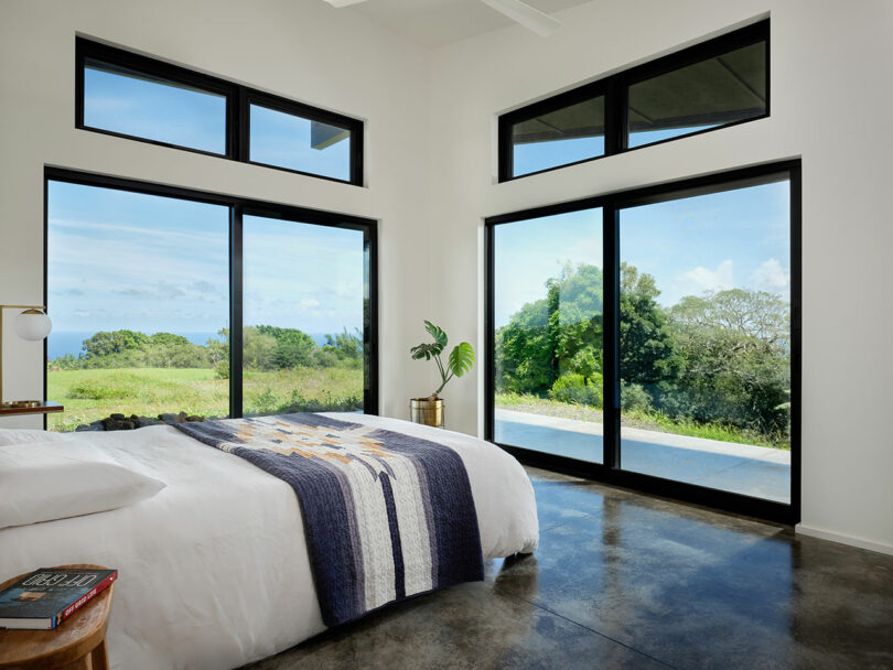 Modern bedroom with large black-framed windows overlooking a green landscape and pool; white bedding with a patterned throw covers the bed.
