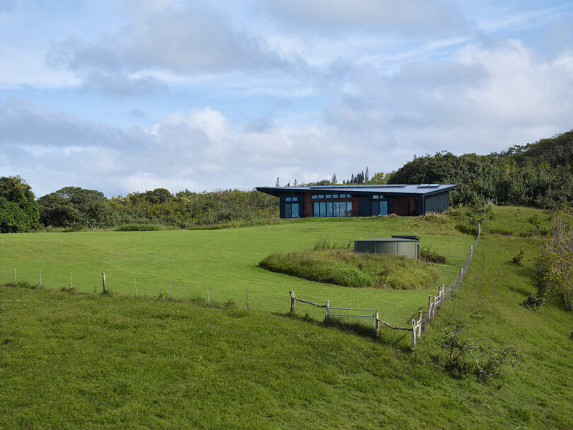 Modern single-story house with large windows sits on a green hilltop, surrounded by grass, trees, and a water tank enclosed by a fence under a partly cloudy sky.