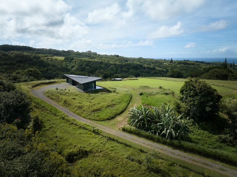 A modern house with a dark roof sits on a grassy hillside surrounded by trees, with a winding gravel road and distant ocean visible under a partly cloudy sky.