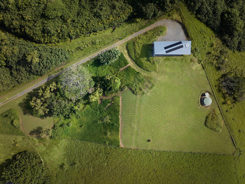 Aerial view of a rural property with a rectangular building, a winding driveway, trees, grassy fields, and a circular water tank.