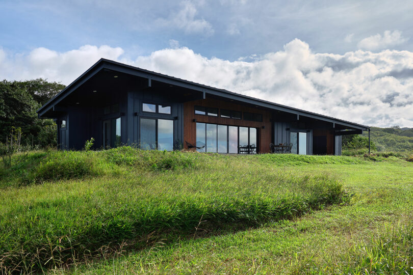Modern single-story house with large windows and a sloped roof, situated on a grassy hill under a partly cloudy sky.