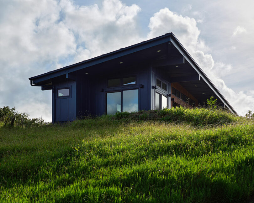 A modern, dark-colored house with large windows sits on a grassy hillside under a partly cloudy sky.