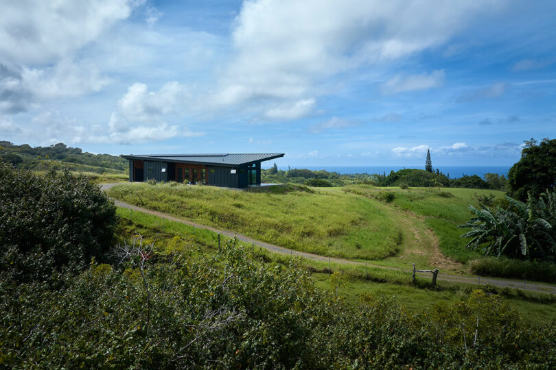 A modern, single-story building with a slanted roof sits in the middle of a grassy landscape under a partly cloudy sky, with the ocean visible in the background.
