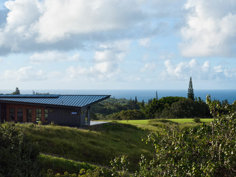 A modern building with a metal roof sits on a grassy hillside overlooking trees and the ocean under a partly cloudy sky.