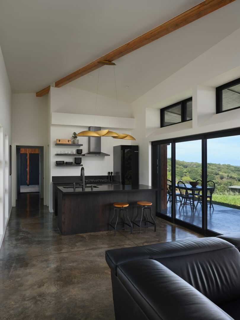 Modern kitchen with dark cabinets, black island, and three stools; large sliding glass doors open to a patio with an outdoor dining set and green landscape visible outside.