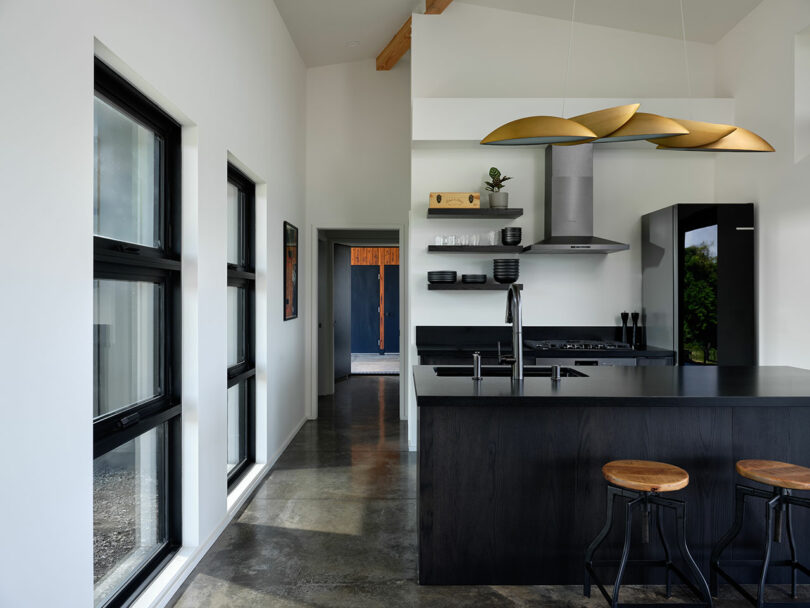 Modern kitchen with black cabinets, island with wooden stools, sleek appliances, open shelving, large windows, and gold pendant lights; polished concrete floor and white walls.