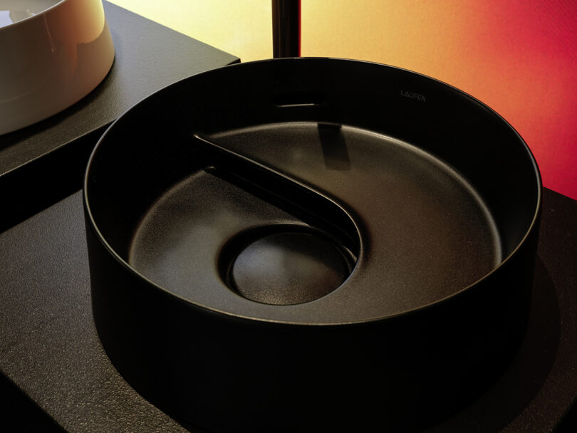 A round, matte black washbasin with a unique spiral divider sits on a dark countertop, illuminated by warm, gradient lighting.