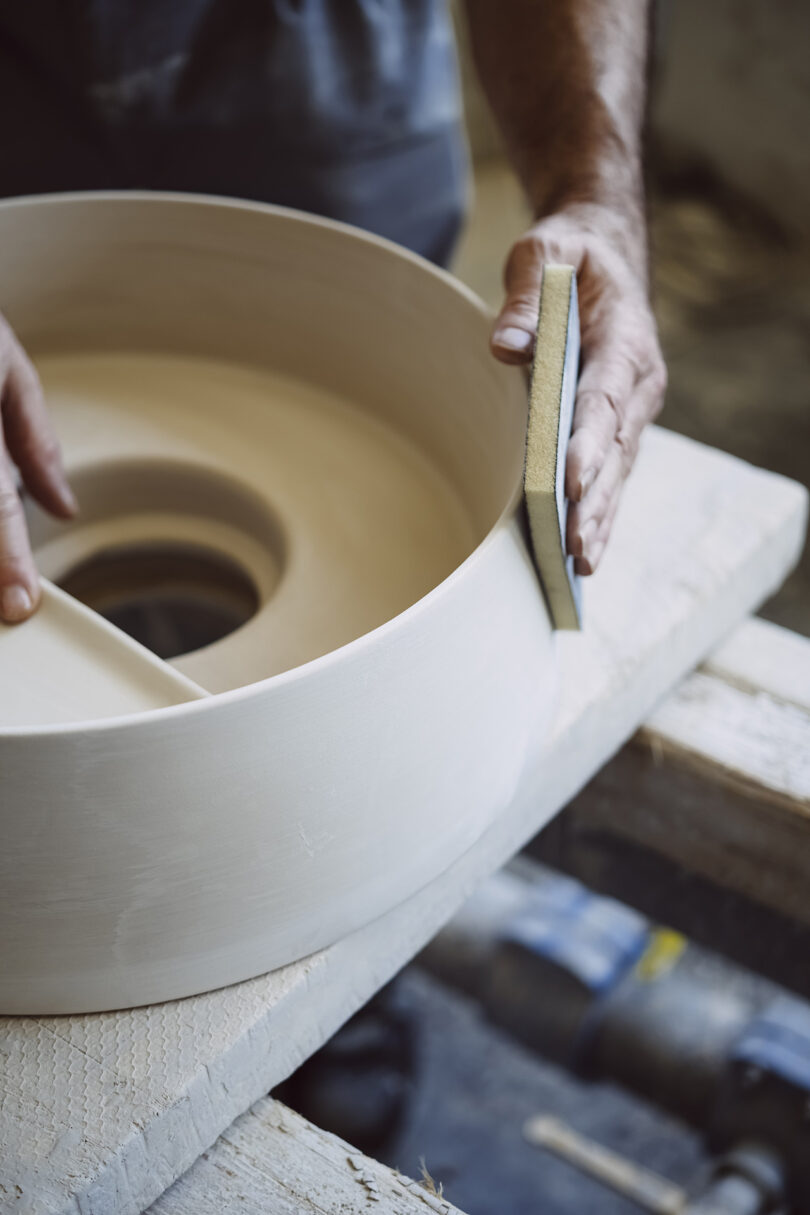 A person uses a sanding sponge to smooth the curved edge of a large ceramic or pottery piece on a wooden workbench.