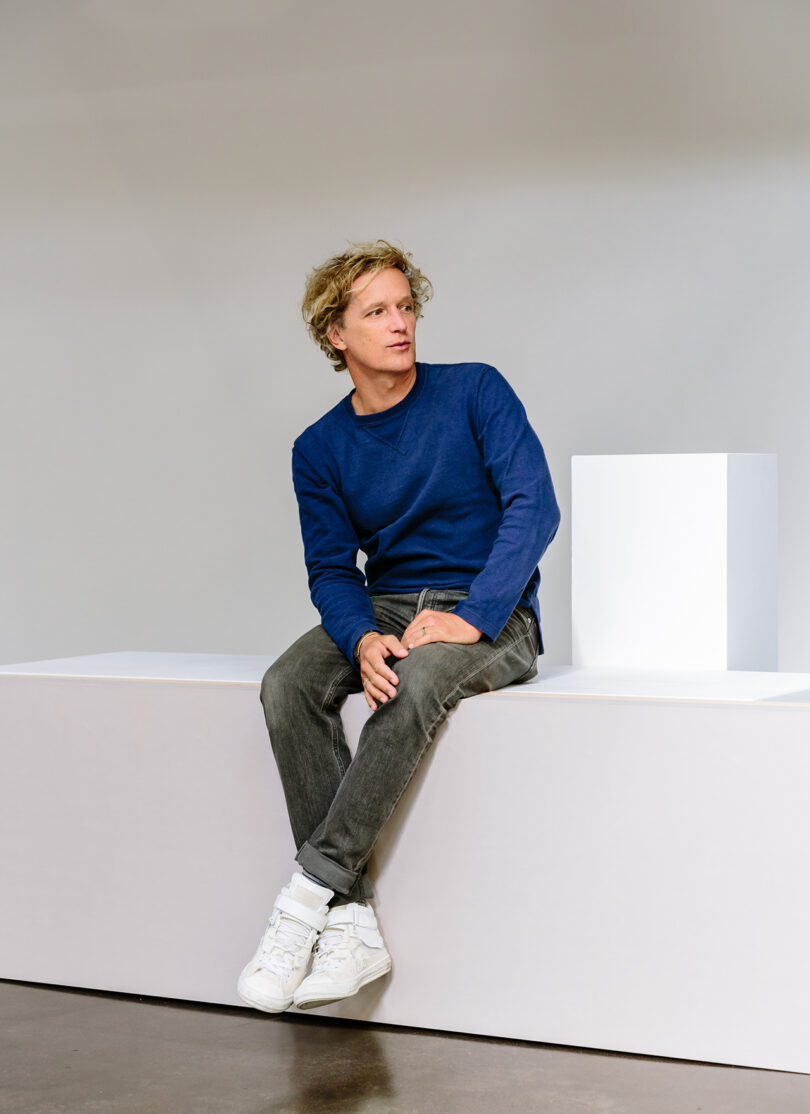 A man with blond hair, wearing a blue long-sleeve shirt, gray jeans, and white sneakers, sits on a white platform against a plain background.