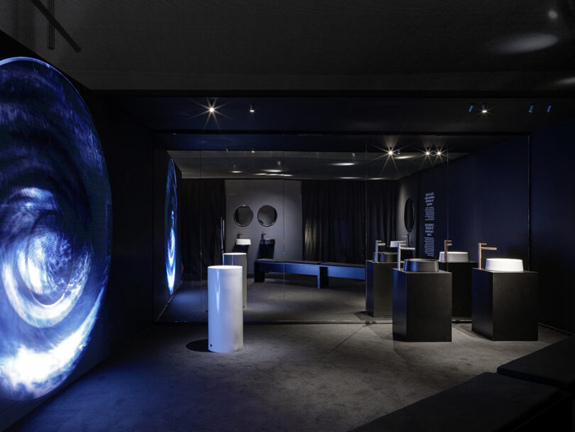 Modern showroom with black walls, illuminated blue circular display on the left, pedestal sinks, mirrors, and minimalistic fixtures arranged in a dimly lit, contemporary space.