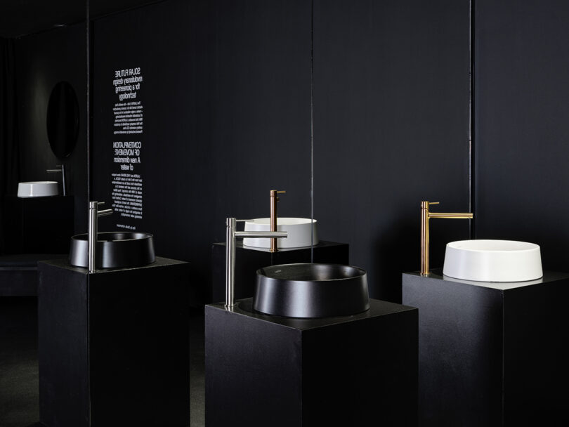 A display of modern black and white round sinks with metallic faucets, arranged on black pedestals in a minimalist showroom with dark walls.