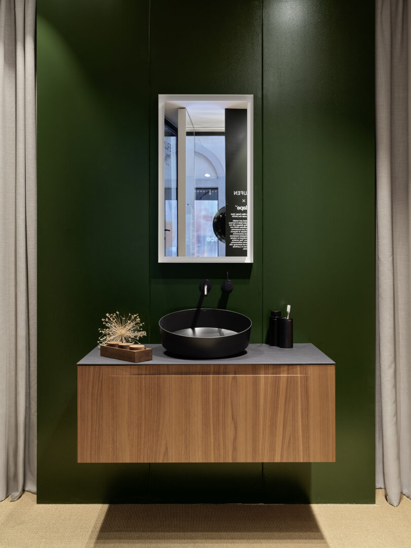 A modern bathroom vanity with a wood finish, a black round vessel sink, a wall-mounted faucet, a framed mirror, and simple accessories against a dark green wall.