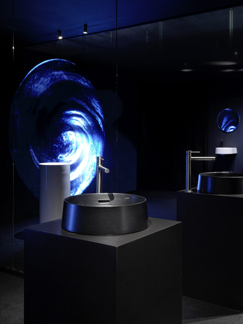 Modern bathroom sinks on black pedestals with a metallic faucet, positioned in a dark room with a blue swirling light projection and a circular mirror in the background.